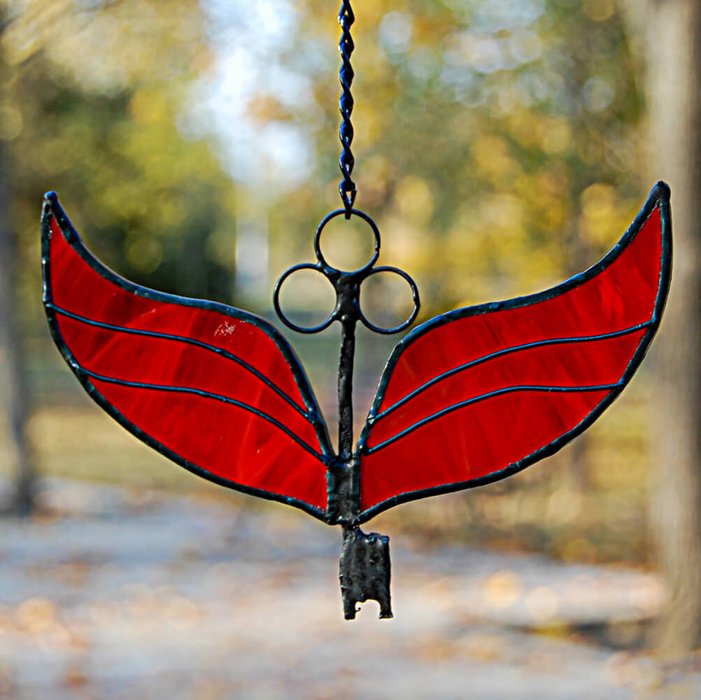 Stained glass red winged key with black wire frame hanging against a blurred natural background