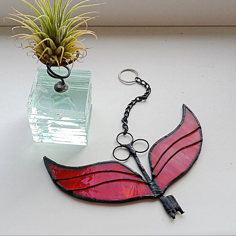 Stained glass butterfly ornament with pink wings and black body on a white surface, next to a small plant in a clear container.